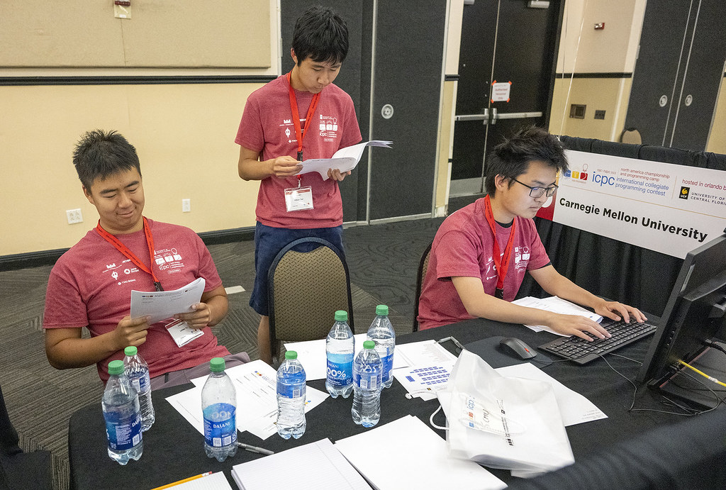  Three male students are in the picture. One sits at a table reading a packet of papers. Another sits at the table in front of a computer. The third is standing behind a chair reading a packet of papers.