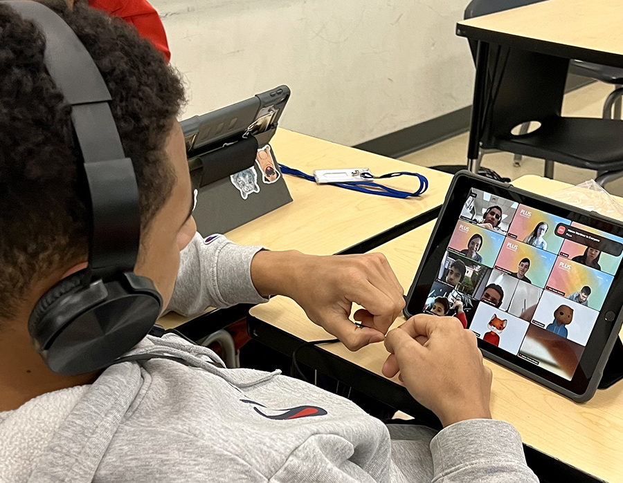  A man with dark hair in a gray hoodie wears headphones and looks at a screen containing a Zoom-like grid of people in 12 squares.of