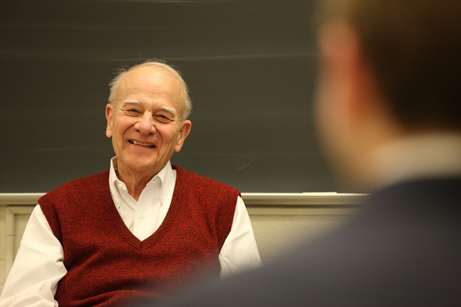  Portrait of William Wulf wearing a red sweater vest over a white dress shirt with a chalkboard in the background and a blurred human shoulder in the foreground.