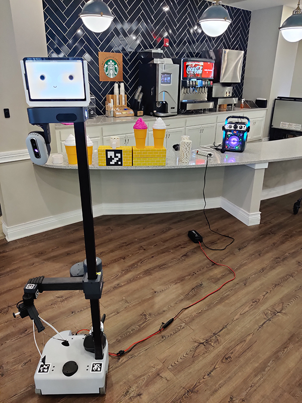  A robot with a touchscreen for a face attached to a pole for a body is positioned in front of an ice cream bar.