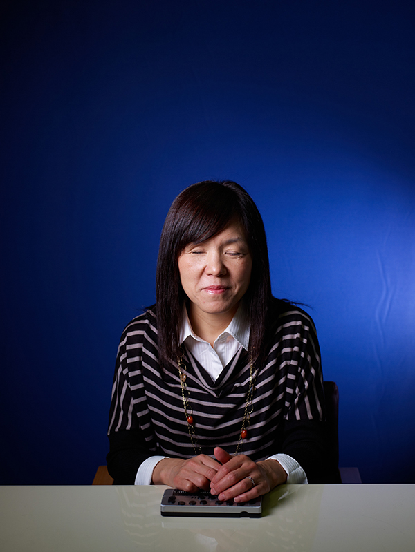  An asian woman sits with her eyes closed at a desk against a blue background with her hands crossed over a small device.