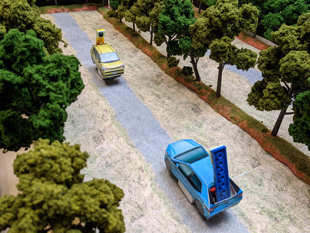  A small green car and a small blue car had toward each other on a board made to look like a one-way street lined with trees.