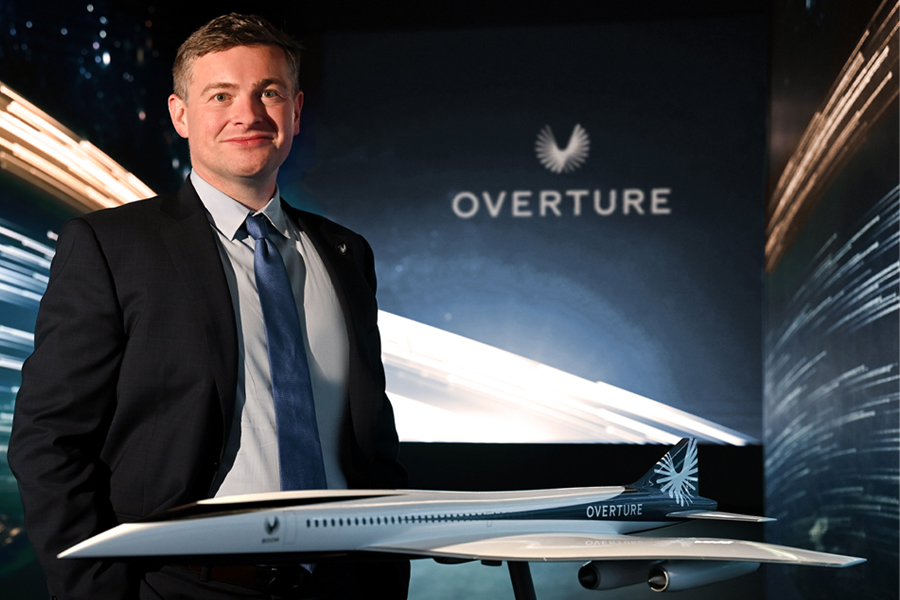  A man stands in front of a dark space-themed background with the word Overture on it, while a model airplane sits in front of him.