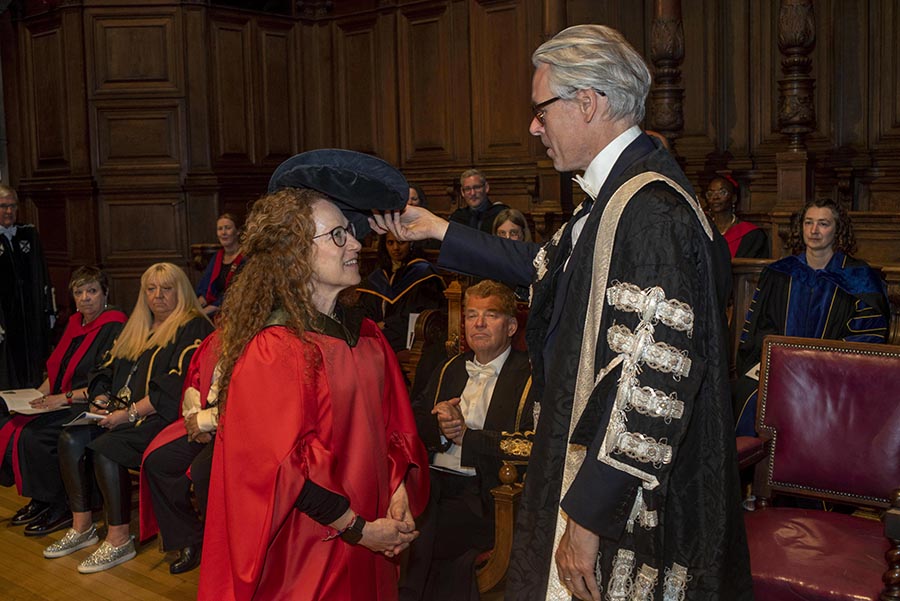  A woman with glasses and curly hair dressed in red academic regalia is tapped on the head with a blue velvet cap called The Geneva Bonnet by a man in black academic regalia.