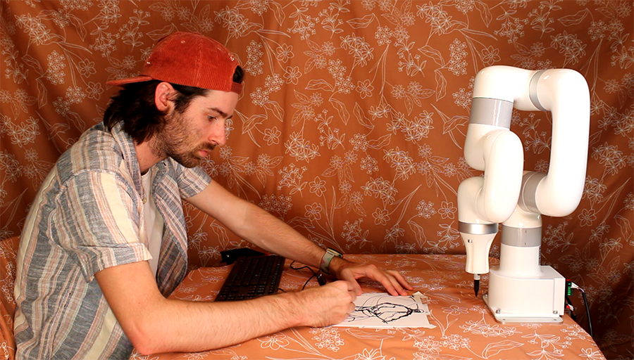  A man wearing a backward baseball cap and striped shirt sits at a table drawing while a robotic arm outfitted with a Sharpie waits across the table for its turn.