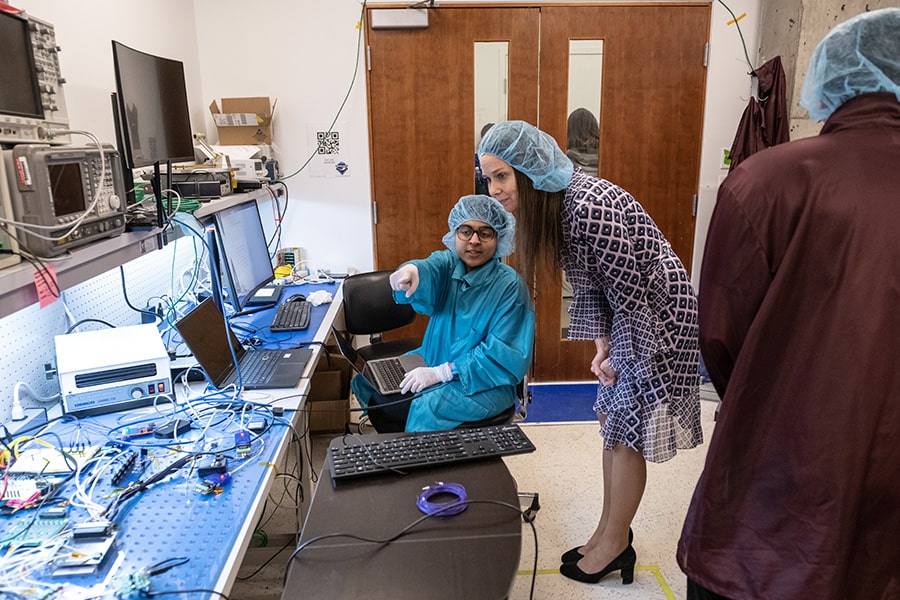  A woman in glasses and clean room garb sits at a desk covered in computing equipment and points at something in the foreground. Another woman, wearing dress clothes and a hair net, bends down to look at what the other woman is pointing at.