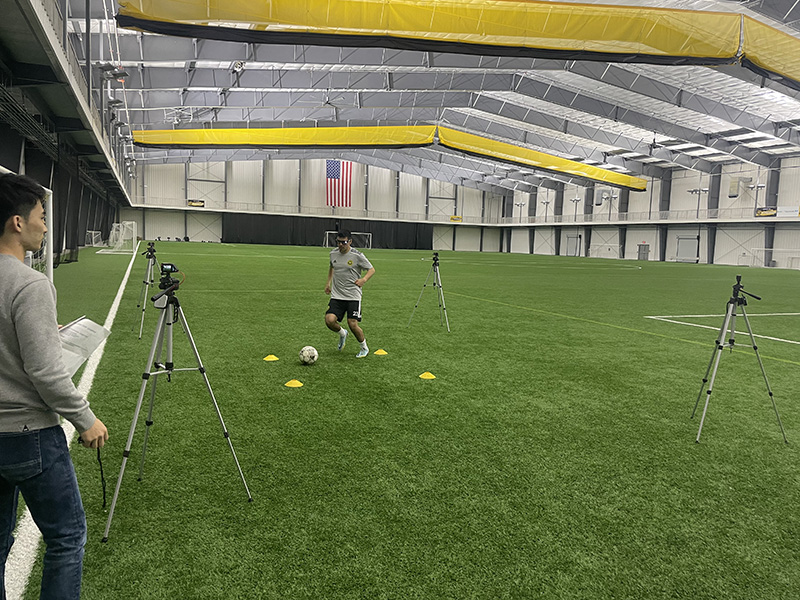  A man surrounded by cameras on tripods kicks a soccer ball while another man with a clipboard looks on.