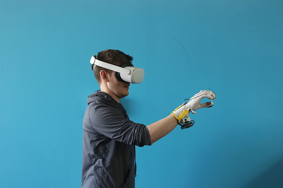  A man in a virtual reality headset stands in front of a teal screen and wears a glove — outfitted with wires and sensors — that conforms to his hand as he makes grasping motions.