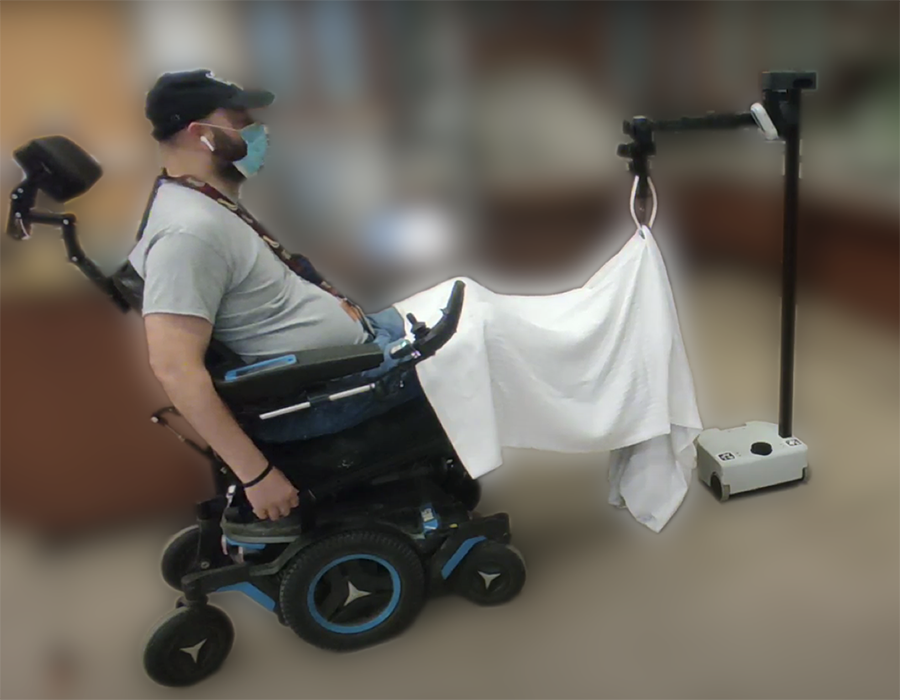  A man in a wheelchair uses a hands-free microphone and head-worn sensor to control a robotic hand that helps him adjust a blanket.