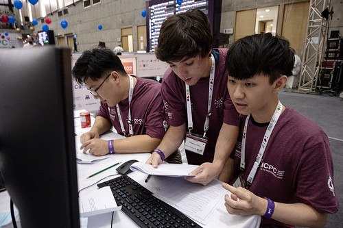  Three students in purple t-shirts sit in front of a computer monitor. One looks at the monitor while two others read papers in front of them.