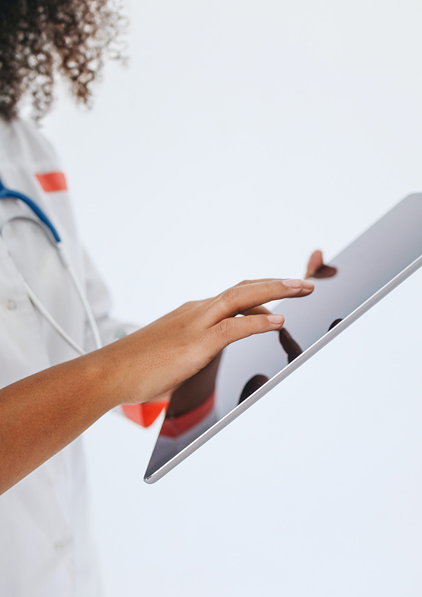  A person with long brown hair in a white coat and stethoscope holds an ipad in their hands.