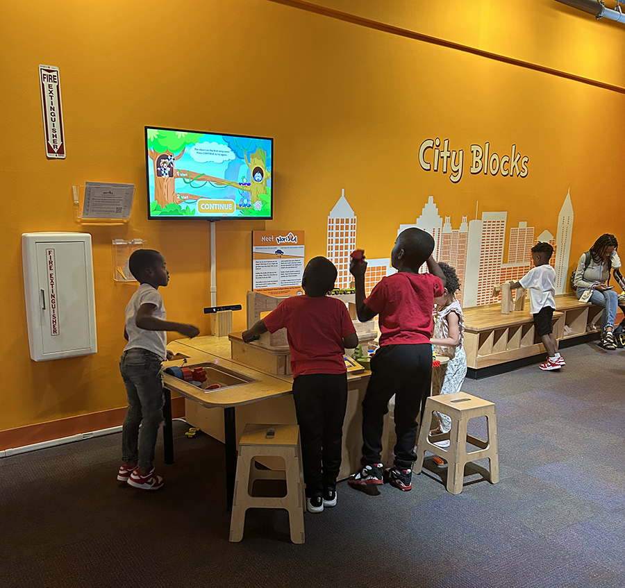  Four children stand around a table covered in blocks while animated characters on a screen behind the table give the children advice for their creations.