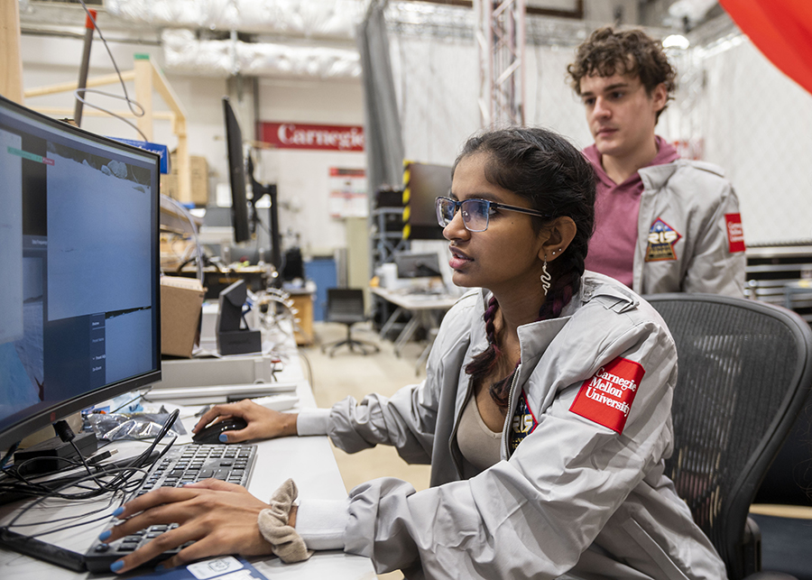  A young woman in a gray jacket with a Carnegie Mellon University patch on the shoulder works at a computer in the foreground while a young man in a matching jacket looks on.