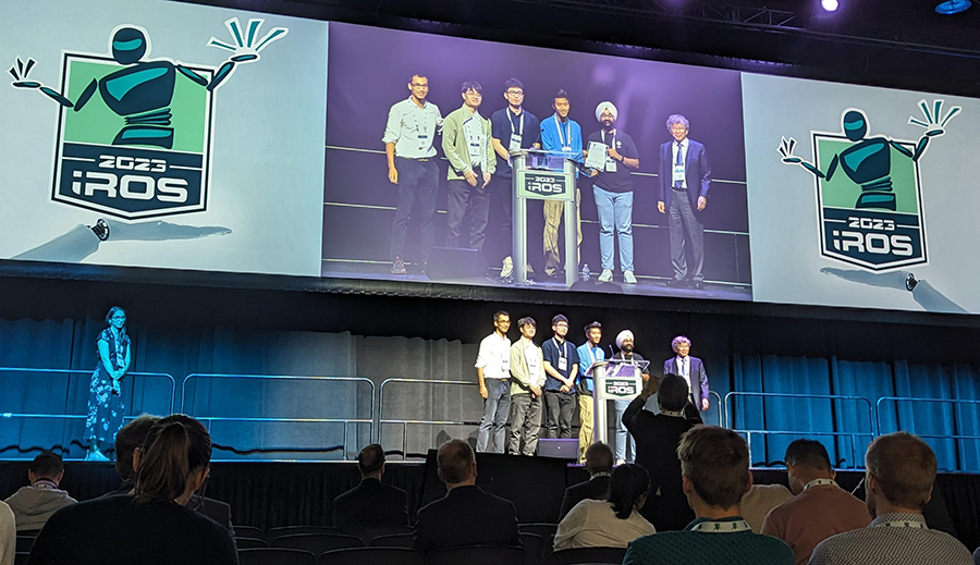  Six men stand at a podium to accept an award with a purple screen and a stylized robot logo behind them.