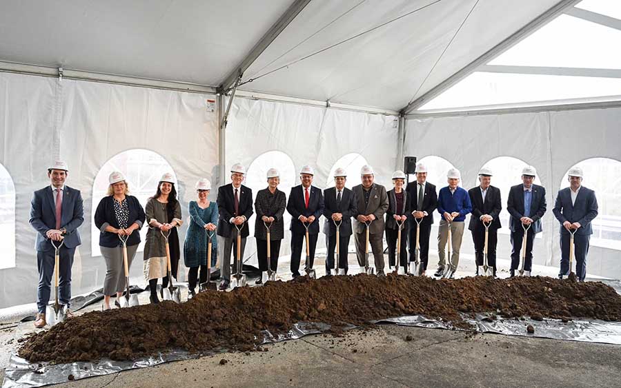  Men and women in various styles of business dress hardhats hold shovels behind a small pile of dirt to commemorate the groundbreaking for a new building.