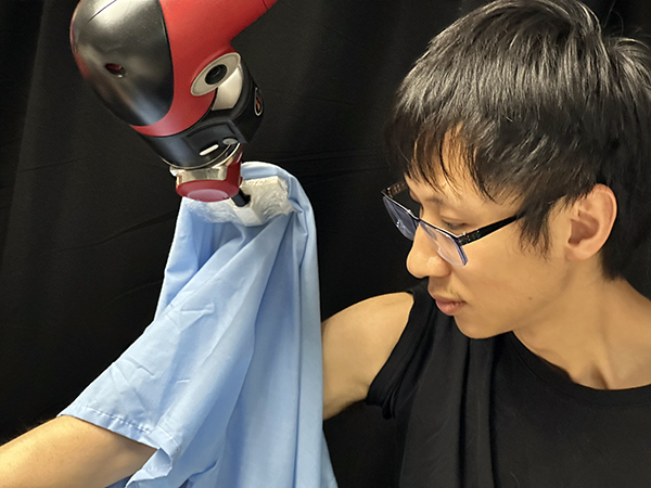  A young man in a black shirt wearing glasses sits in front of a black backdrop while a roboti arm slides a blue shirt sleeve up his arm.