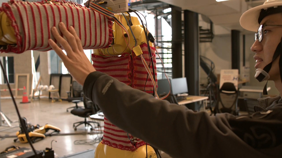  A male on the right side of the photo wearing glasses and a hard hat touches a robotic arm on the right that is covered in red and white knitted fabric.