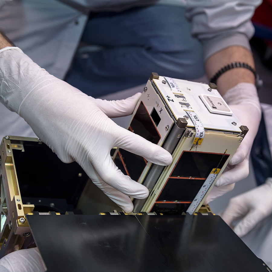  A pair of human hands in white gloves manipulate a small box-shaped satellite, preparing to place it into a box with three other identical satellites..