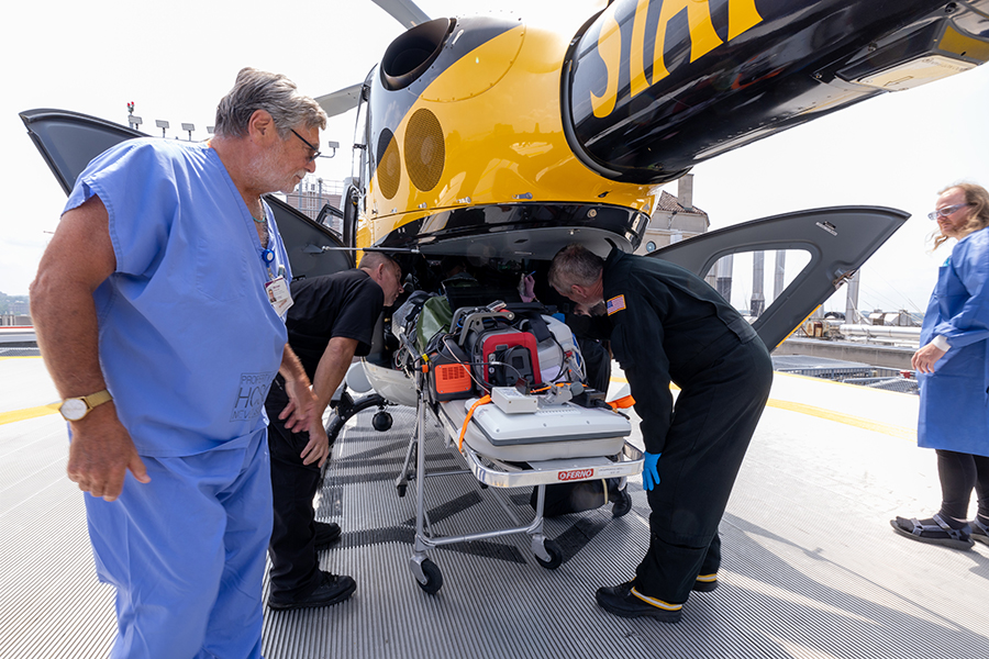  A man in blue scrubs stands near a medical helicopter on a helipad while two medics dressed in black inspect a gurney outfitted with an autonmous trauma care system.